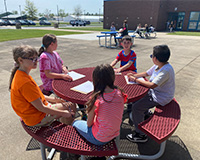 A group of five students sitting around an outside table taking notes on paper